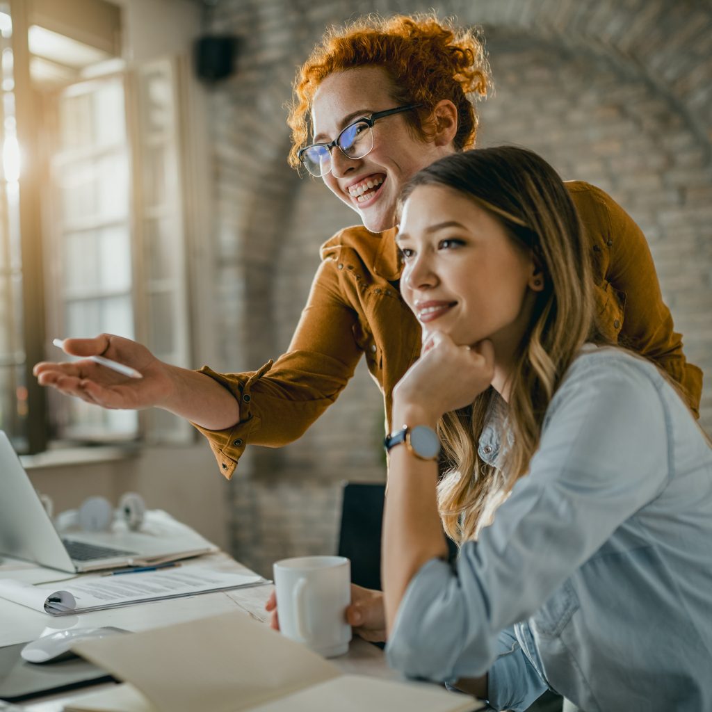 Two happy female entrepreneurs, including a redhead woman, reading an email on the computer while working together in an office for Maked.